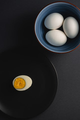 Boiled egg with yellow yolk in black plate near to bowl with white eggs on dark moody black plain minimal background, angle view, happy Easter day