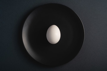 Single white egg in black plate on dark moody plain minimal background, top view, happy Easter day