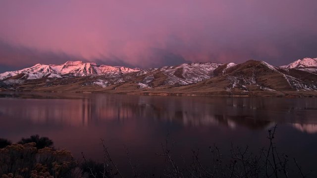 Time lapse of low clouds lighting up as sun rises and shines on snow covered mountain range reflecting in lake in Utah.