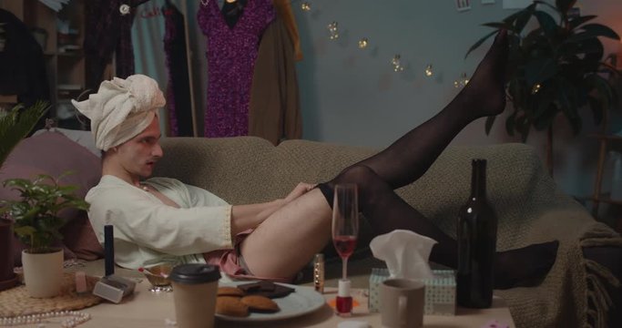 Man transgender getting ready for date while lying on sofa and trying to put on black tights. Home background. Concept of people, beauty and leisure.