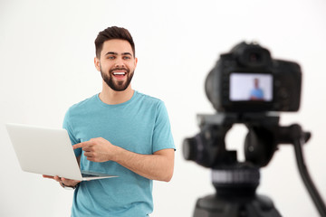 Young blogger with laptop recording video on camera against white background