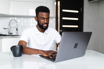 Smiling african american man drinking coffee and using laptop on the kitchen