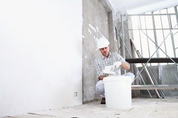 plasterer man at work, take the mortar from the bucket to plastering the wall of interior construction site wear helmet and protective gloves, ladder and scaffolding on background © amedeoemaja