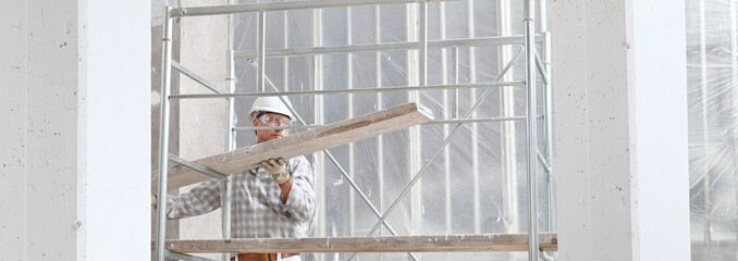 man work, professional construction worker on scaffolding safety hard hat, gloves and protective glasses. on interior building site background, panoramic image