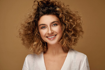 Happy young woman with healthy natural curls and delicate make-up posing at studio on brown studio background. Blonde woman with afro hairstyle smiling