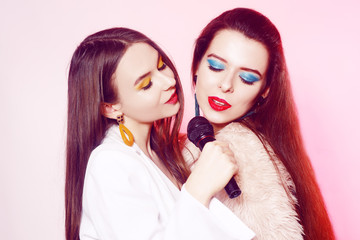 Beautiful brunettes on a pink background sing into a microphone. Teenager in karaoke. Girl with bright green make-up, earrings and red lips. Sisters on a pink