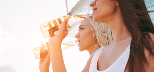 Closeup of two attractive cheerful girls friends in stylish swimsuits drinking beer and having fun on sunny beach. Beautiful young women posing with bottles of beer. Summer travel at seaside