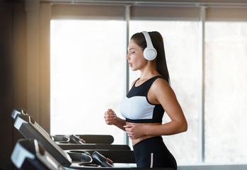 Pretty young woman with headphones listening to music while running on treadmill in gym