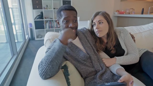 Mid shot of a multiracial couple playfully fighting over the remote whilst cuddling on the couch in front of the TV. 