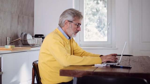 Slow motion shot of mature man using laptop in the kitchen and typing