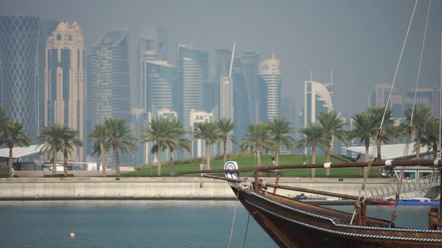 Retro historical boat with blurred panoramic view of modern skyline of Doha and green palms on background