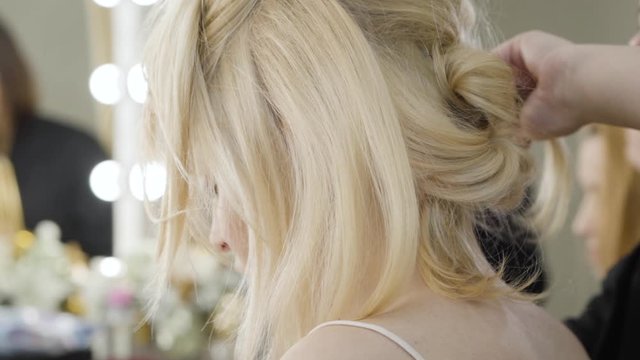 Close-up of hairdress of blond young woman sitting in beauty salon. Beautiful rich Caucasian lady doing hairstyle. Stylist working with client. Style, fashion, beauty. Slow motion.