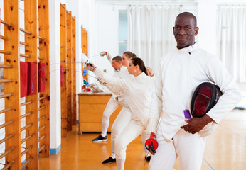 Active african american male fencer in uniform standing with mask and foil at fencing room
