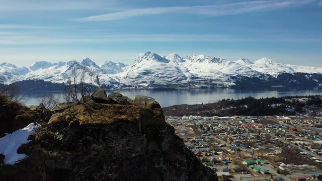 Flying over the edge of a mountain cliff to reveal the city of Valdez, Alaska and its snowy surroundings in April.