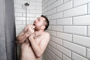 Shocked man looks at a watering can in the shower room, from which, unexpectedly, cold water is pouring. - obrazy, fototapety, plakaty
