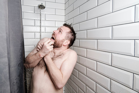 Shocked man looks at a watering can in the shower room, from which, unexpectedly, cold water is pouring.