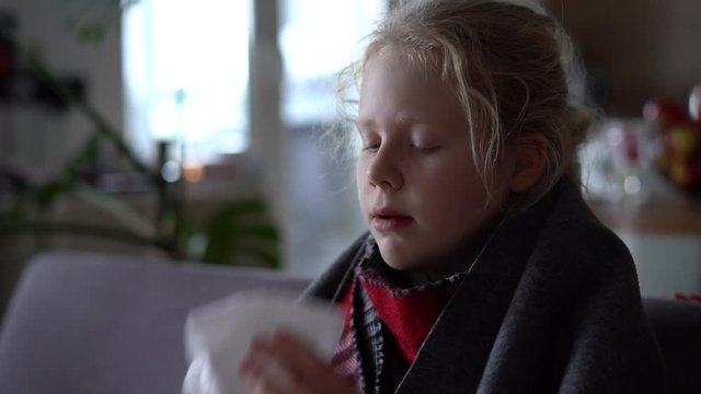 cold caucasian girl at home. portrait of a sick child in a scarf and plaid on the sofa in the apartment, the schoolgirl blows snot into a napkin