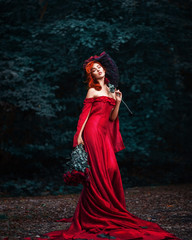 Amazing beautiful cute young woman with fiery red hair in red long dress stands on path in green forest with bouquet of red roses in her hands.