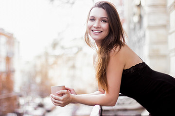 Portrait of happy young woman in black pajamas holding cup of coffee in hands and smiling while standing on balcony. Beautiful gentle french girl in dress drinking hot tea and smiling on home terrace