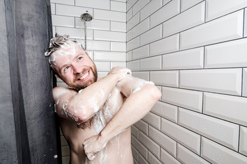 Cute, happy man soaped in foam washes in the shower, under running water, he smiles and looks dreamily to the side.