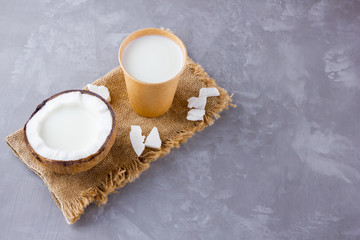 Obraz na płótnie Canvas Coconut milk and coconut in paper glass on gray background. Coconut vegan milk non dairy on sackcloth. Healthy drink concept. Alternative milk. Top view