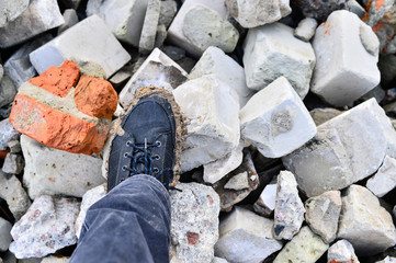 A man's foot in a dirty leather boot stands on a pile of crushed bricks.