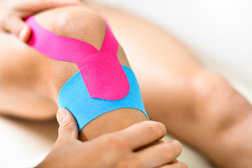Kinesiology taping treatment with blue and pink tape on athlete patient injured knee. Woman hands...