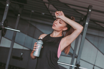 Exhausted attractive sports woman holding bottle of water in hand and relaxing while standing in...