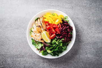 Healthy salad buddha bowl plate with cherry tomatoes, chicken, beans, sweet pepper, lime and mixed greens on grey background top view. Healthy food. Superfood meal