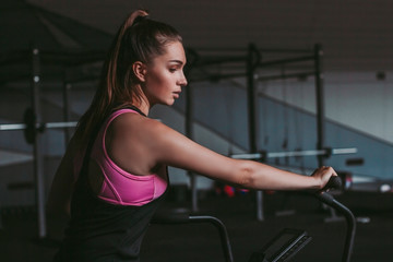 Fototapeta na wymiar Portrait of beautiful determined fitness woman in sports wear doing cycling exercises in dark gym. Attractive slender sports girl exercising on bicycle indoors. Intense functional training in gym