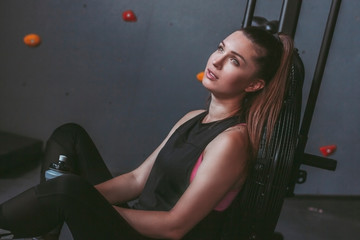 Portrait of exhausted beautiful sports woman sitting on floor and holding bottle of water in hands while relaxing after cycling training in gym. Tired fitness girl having rest after workout on bicycle