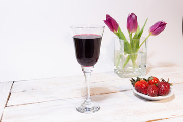 glass of red wine, strawberry on plate, pink tulips in vase on white wooden background