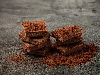 Broken pieces of chocolate sprinkled with coffee powder close up on gray stone background.