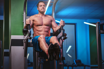 Fototapeta na wymiar Powerful athletic bodybuilder doing excersise on a parallel bars, pumping his abs and lifting his legs in a modern fitness center looking diligent