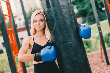 Portrait of attractive young fitness woman holding boxing bag after training outdoors. Beautiful blonde sports girl in boxing gloves hugging punching bag while posing on sports ground. Martial arts