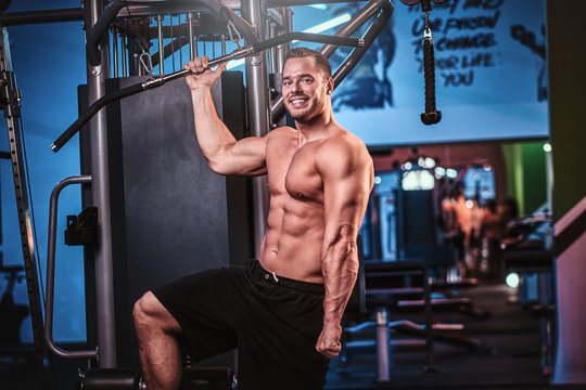 Athletic shirtless man posing for a camera while holding a bar, standing next to back pulling machine in a modern fitness club looking cheerfull
