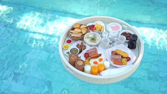 Top view on breakfast food tray floating on pool water. Cofffe cups, glasses with juice, bread, fruits, eggs bacon of floating tray