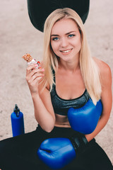 Portrait happy young fitness woman holding protein bar in hand and smiling while sitting at sports...