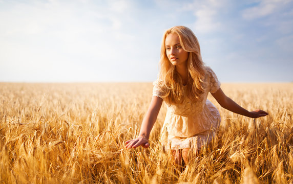Girl with blond wavy hair in white lace dress, posing on wheat field and looking away. Blurred background blue sky with white clouds