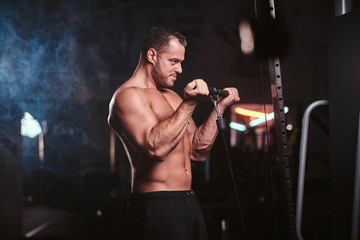 Fototapeta na wymiar Powerful adult bodybuilder pumping up his biceps on a hand pull machine in a dark gym under the spotlights surrounded by smoke looking tensed