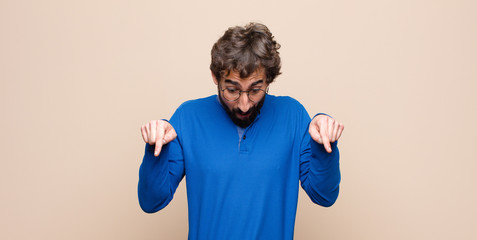 young handsome man with open mouth pointing downwards with both hands, looking shocked, amazed and...