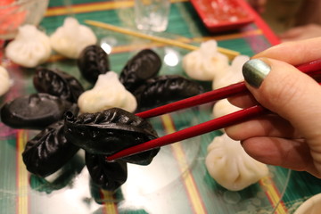 Two red sticks in a woman's hand hold a black dim sum over a dish with other dim sum. 