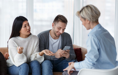 Wife Talking With Psychologist While Husband Using Smartphone In Office