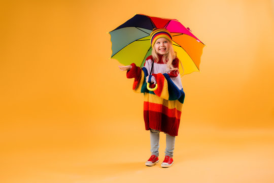 child girl smiling in a bright multicolored sweater and hat holding an umbrella stands on a yellow background. little girl in bright clothes is holding a red-colored umbrella. happy child