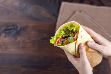 Burrito with slices of fried beef, tomato, lettuce, wrapped in cheese tortilla in female hands. Mexican national cuisine. Top view on food on the background of a wooden table with copyspace.