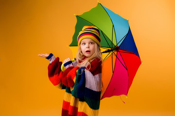 cute little girl in a knitted sweater and hat is holding a multicolored umbrella on a yellow...