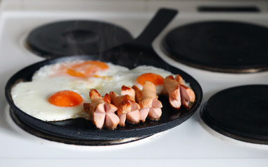 Fried eggs with sausages in iron pan at white electircal cooker