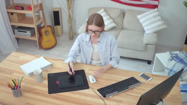 Creative Girl Creates Design On Graphic Tablet, Young Designer Lady In Glasses Works With Gadget And Computer While Sitting At Wooden Desk In Stylish Home Interior, Top View