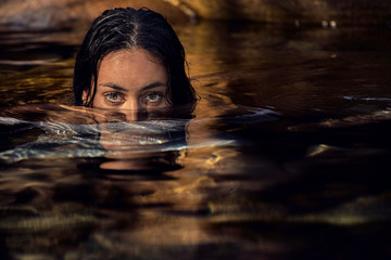Joven mujer morena con la mitad de su rostro y su cuerpo sumergido en el agua mirando hacia adelante con sus hermosos ojos - obrazy, fototapety, plakaty