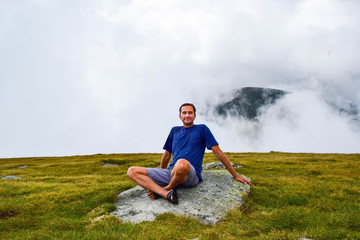 The man enjoys the sun's rays, sitting on a stone surrounded by fog and low clouds near Transalpina...